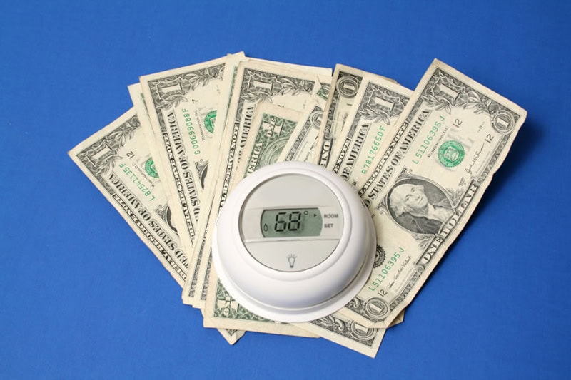 Spring Money-Saving Tips. Photo of a thermostat on top of dollar bills, laying on a blue table or flooring.